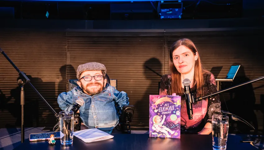 Raúl Krauthausen und Adina Hermann an einem Tisch mit befestigten Mikrofonen sitzend. Auf dem Tisch ihr Buch.