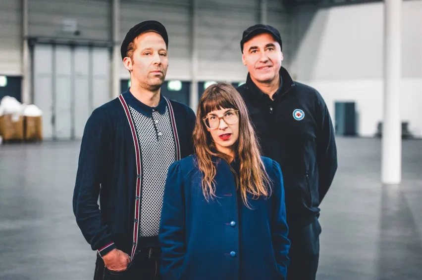 Drei Personen stehen in einer Halle. Im Vordergrund eine Person mit langem Haar und blauem Mantel, hinter ihr zwei, die jeweils eine Kappe tragen.