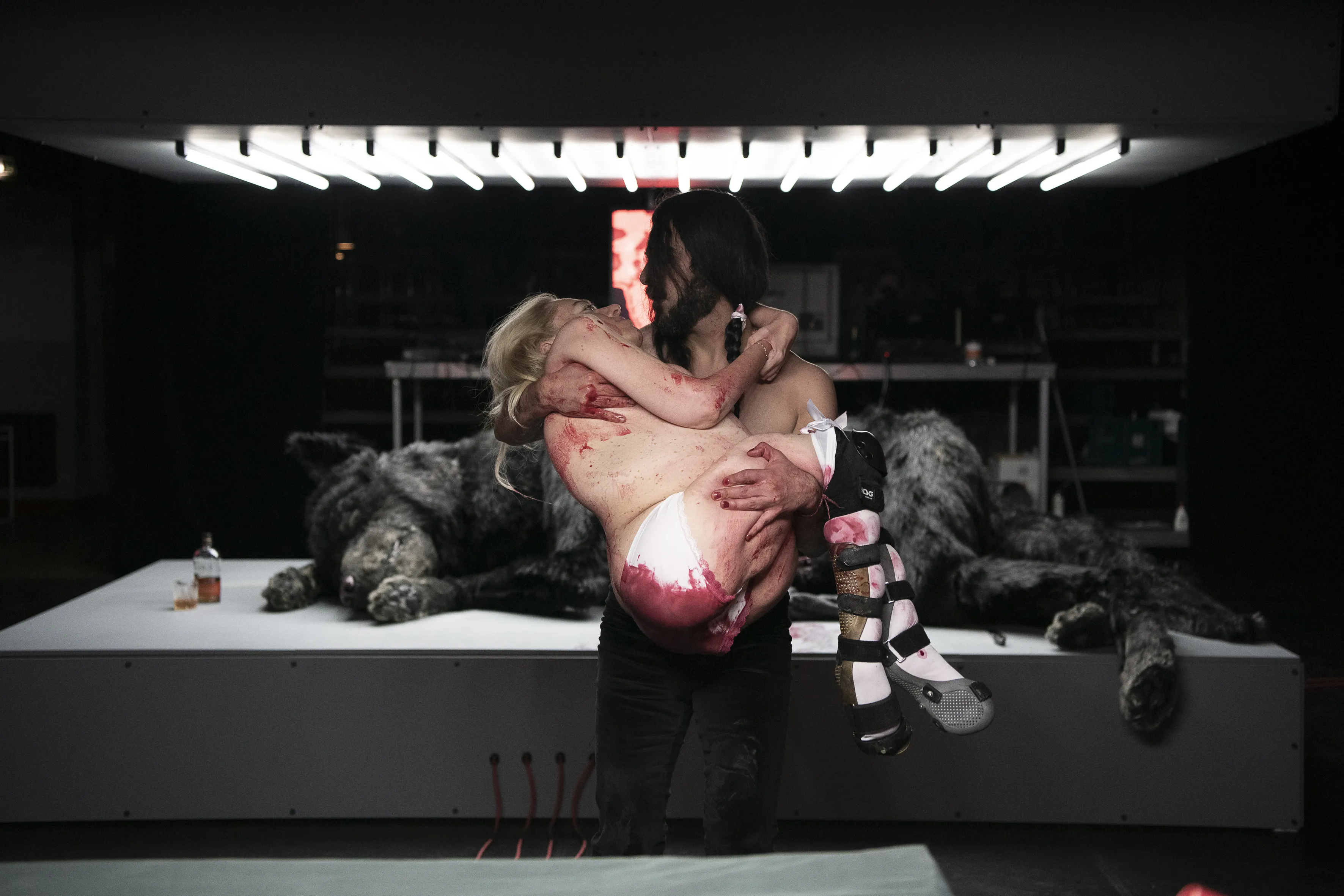 Eine Person, die eine andere, fast unbekleidete Person mit roten Flecken in den Armen hält. Im Hintergrund liegt ein schwarzer Wolf auf einer beleuchteten Fläche.