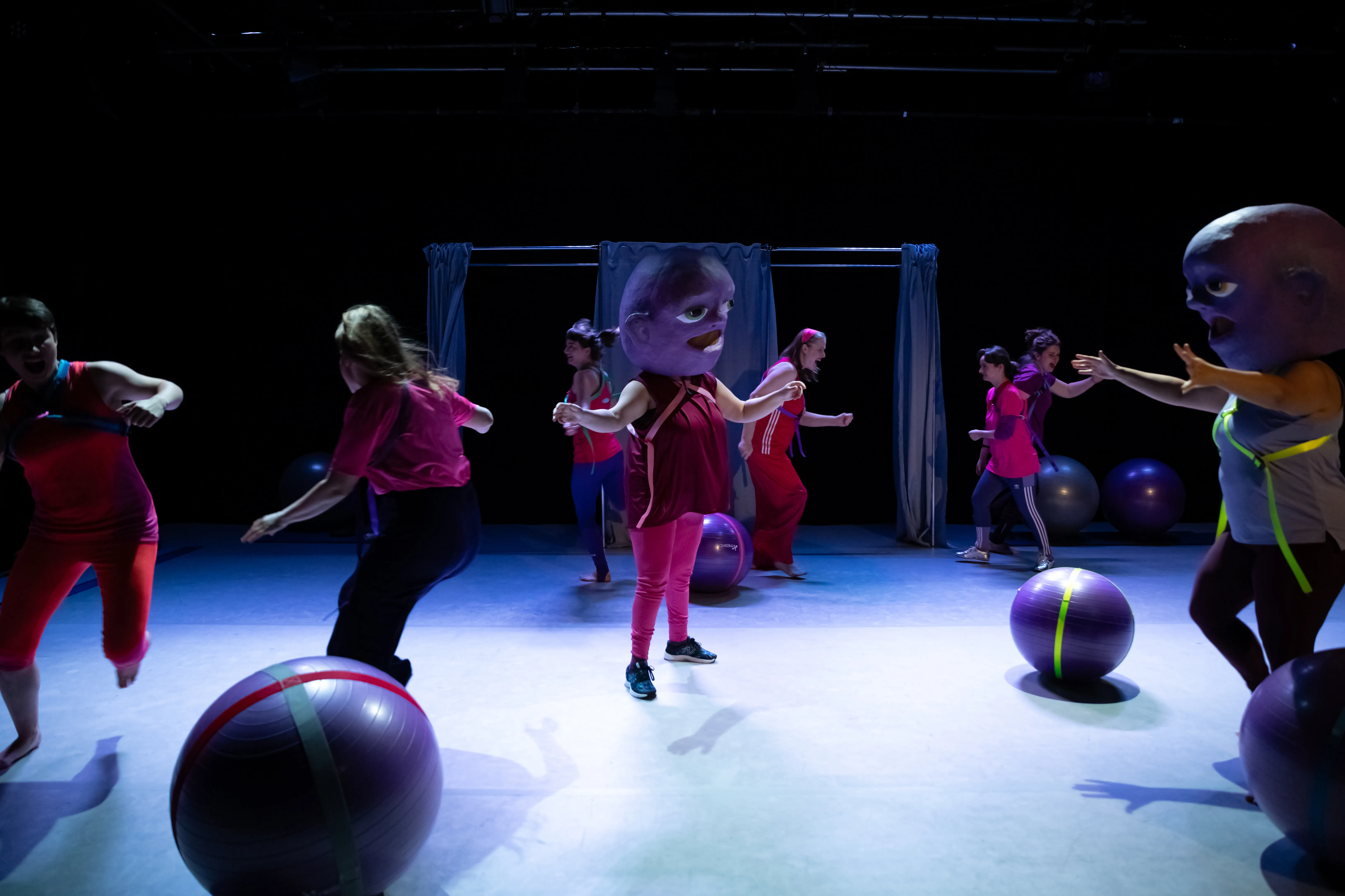 Acht Personen bewegen sich um Gymnastikbälle. Zwei von ihnen haben eine große Maske auf dem Kopf.