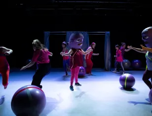 Acht Personen bewegen sich um Gymnastikbälle. Zwei von ihnen haben eine große Maske auf dem Kopf.