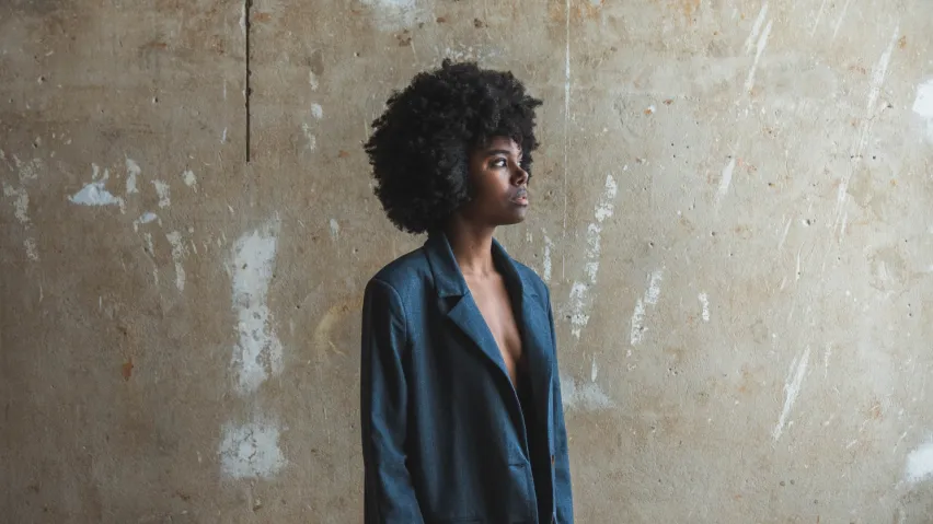 Eine Person trägt einen dunkelblauen Blazer und steht vor einer hellen Wand.