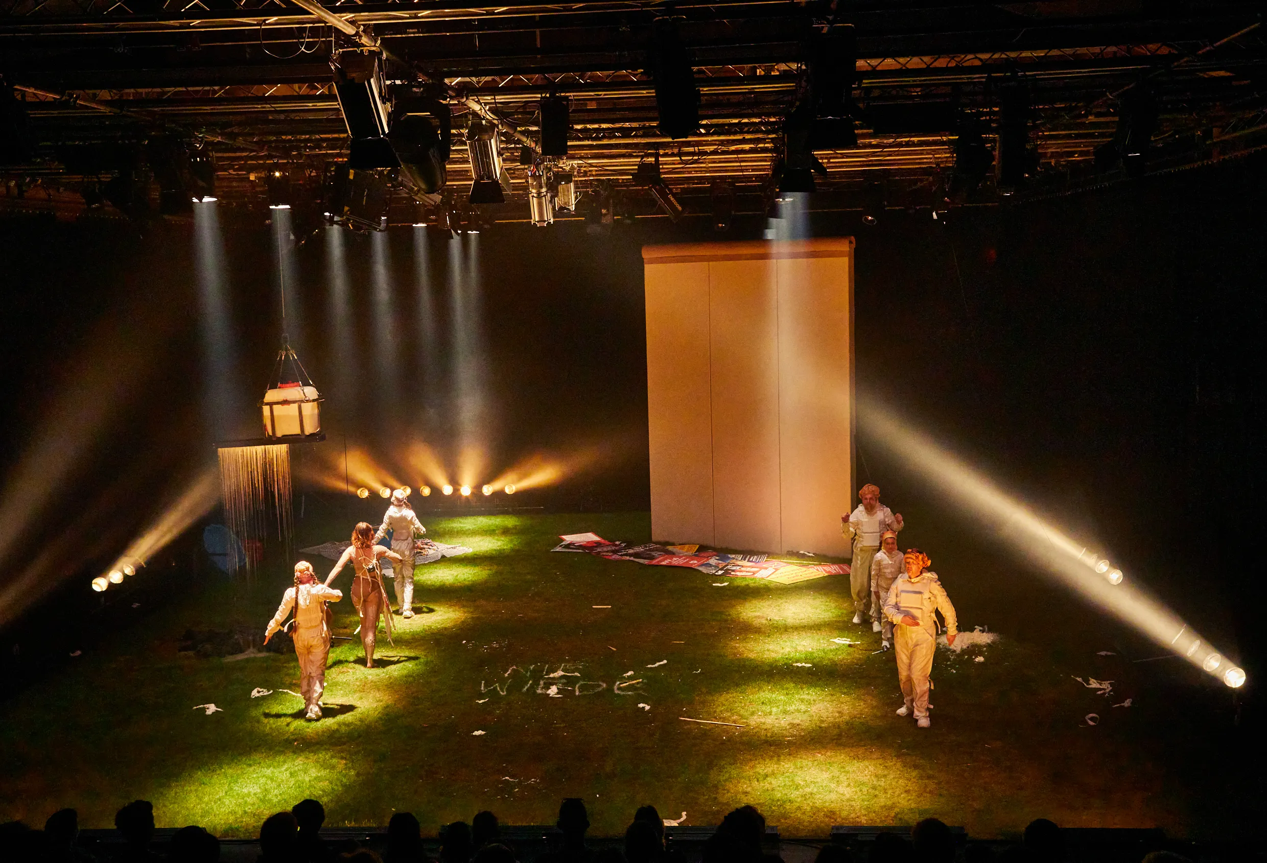 Bühnenbild mit grünem Boden, sechs Personen in hellen Kostümen, Scheinwerferlicht und einer großen weißen Wand im Hintergrund.