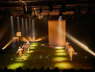 Bühnenbild mit grünem Boden, sechs Personen in hellen Kostümen, Scheinwerferlicht und einer großen weißen Wand im Hintergrund.