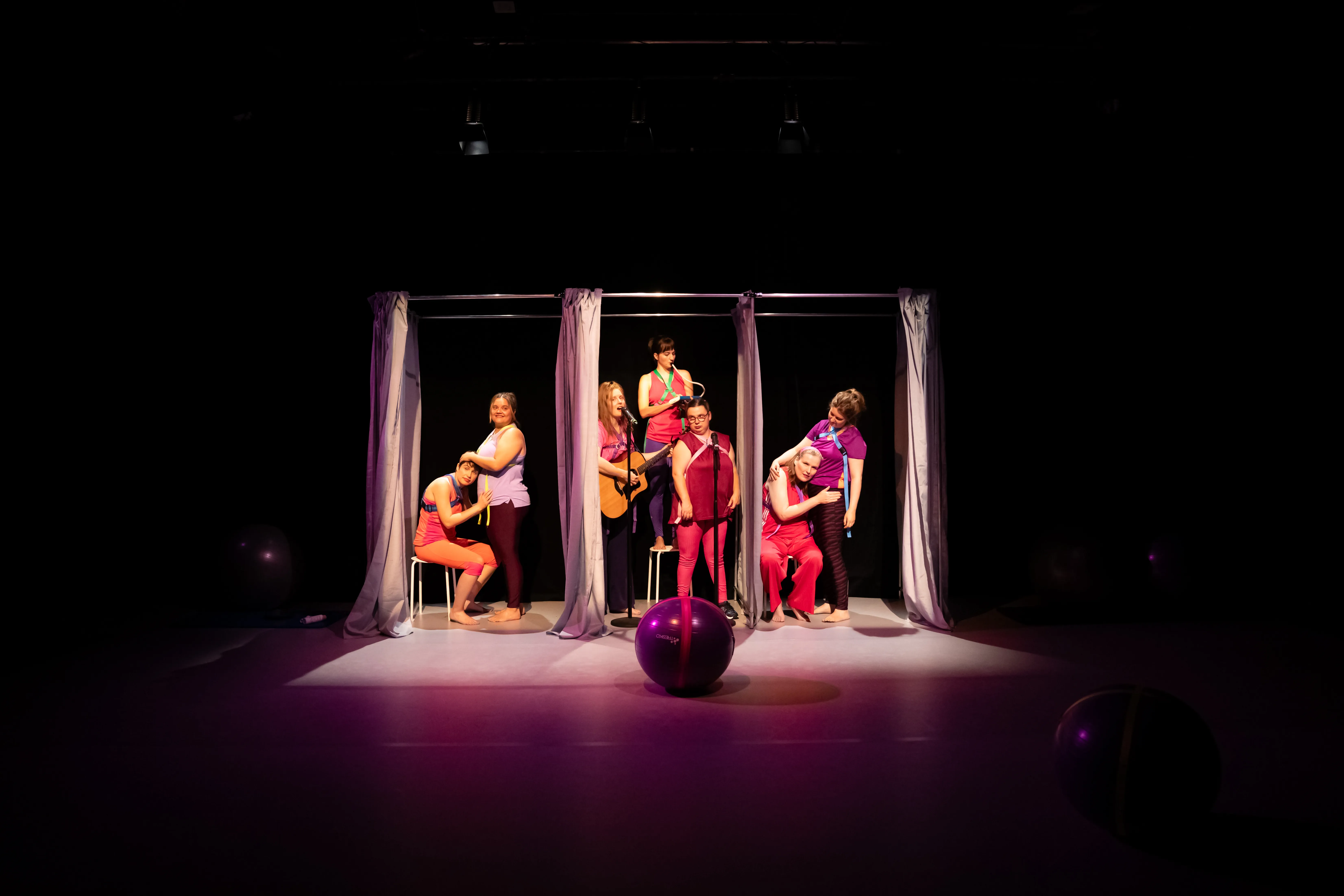 Ein Gymnastikball liegt vor sieben Personen, die sich zwischen Gardinen befinden. Eine von ihnen steht mit Gitarre vor einem Mikrofon.