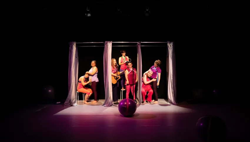 Ein Gymnastikball liegt vor sieben Personen, die sich zwischen Gardinen befinden. Eine von ihnen steht mit Gitarre vor einem Mikrofon.