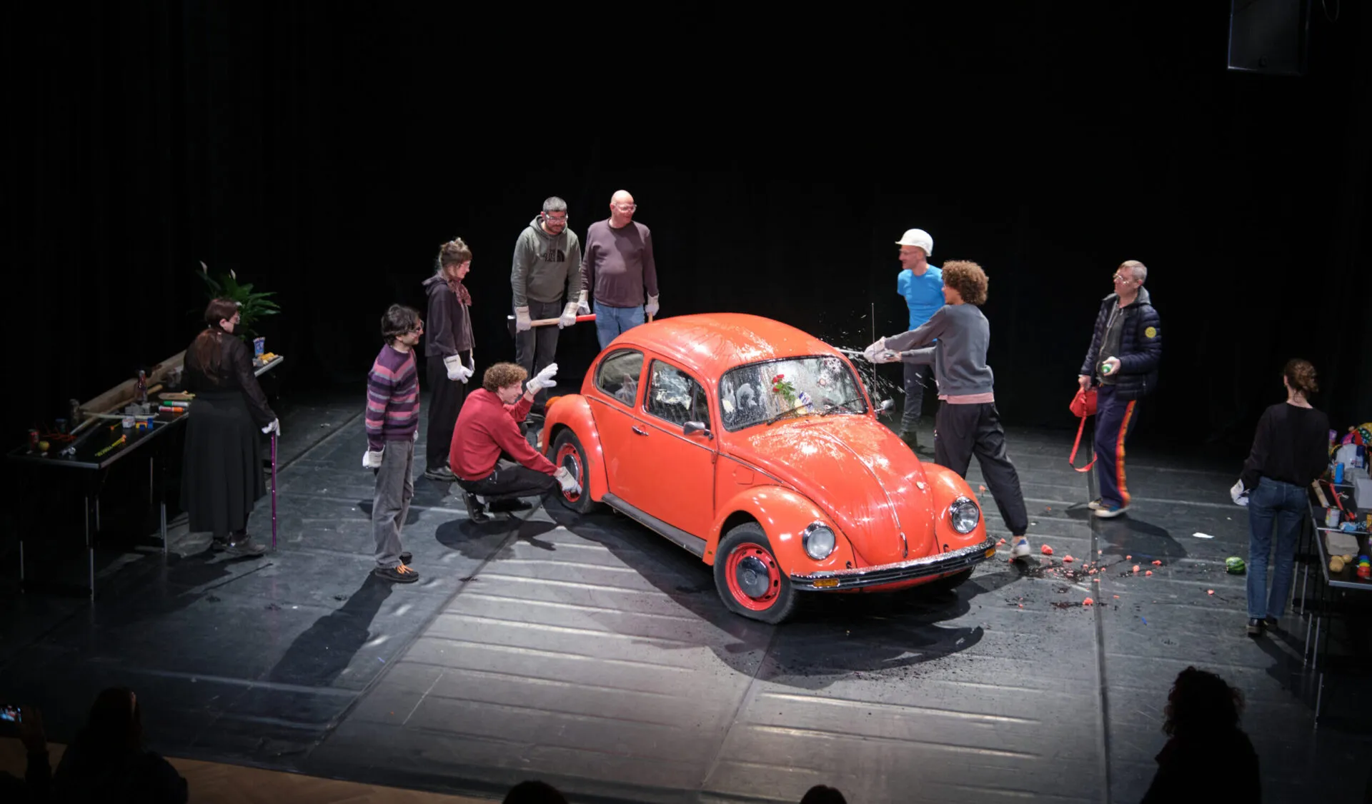 Ein roter VW Käfer, umgeben von mehreren Personen. Eine davon schlägt die Scheibe ein.