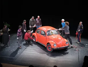 Ein roter VW Käfer, umgeben von mehreren Personen. Eine davon schlägt die Scheibe ein.