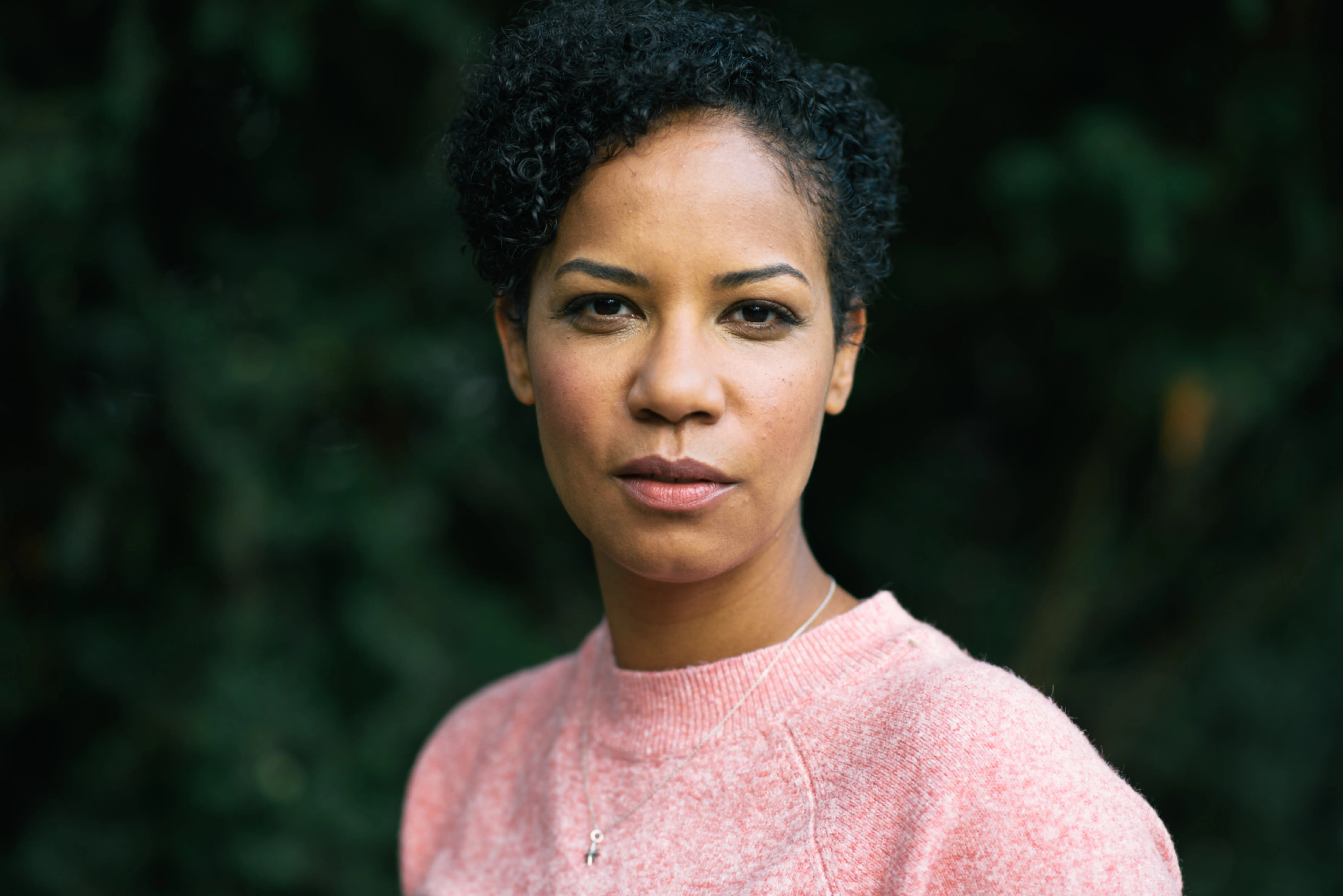 Porträtbild einer Person in einem rosa Pullover. Verschwommenes Grün im Hintergrund.