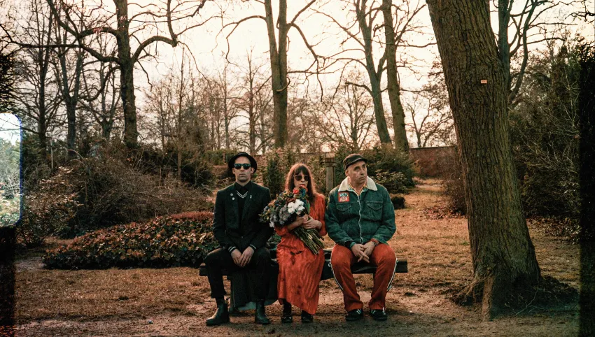 Drei Personen sitzen auf einer Bank. Die mittlere Person hält einen Blumenstrauß.