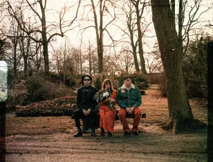 Drei Personen sitzen auf einer Bank. Die mittlere Person hält einen Blumenstrauß.