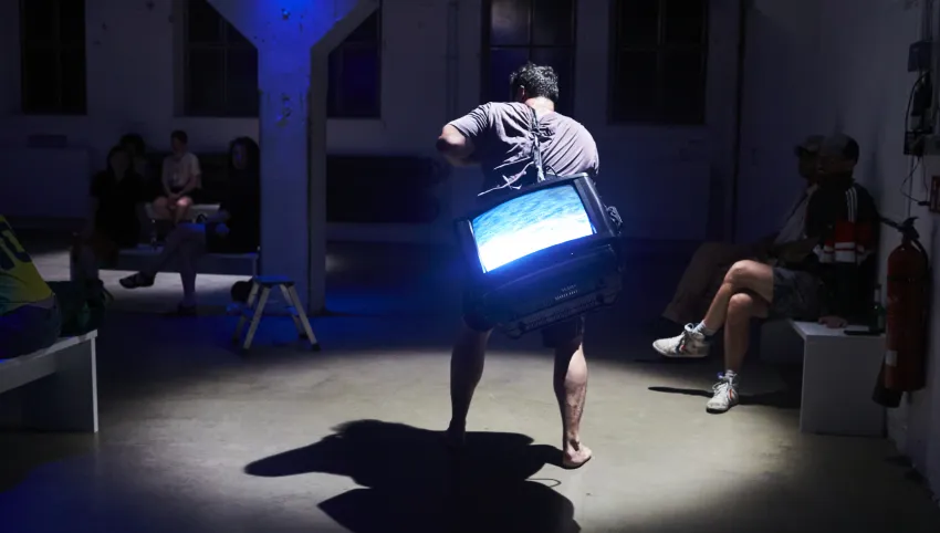Eine Person, die einen Fernseher auf ihrem Rücken trägt und barfuß durch einen Raum läuft. An der Seite sitzen andere Menschen.