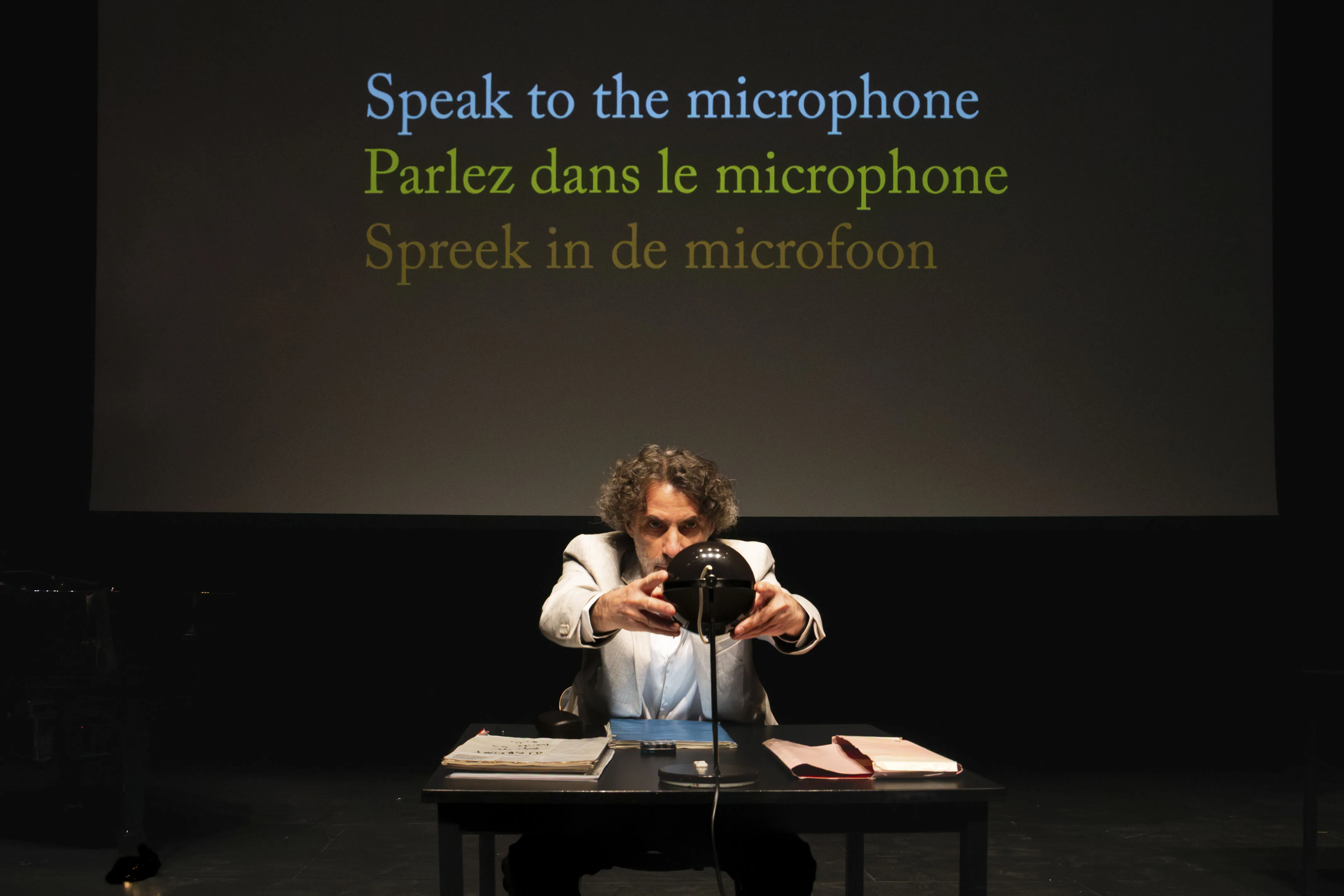 Eine Person an einem Tisch. Hinter ihr steht „Speak to the microphone. Parlez dans le microphone. Spreek in de microfoon.”