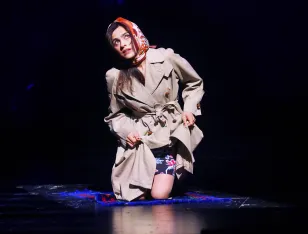 Eine Frau kniet und blickt nach oben. Sie trägt einen beigen Mantel und ein rotes Kopftuch.