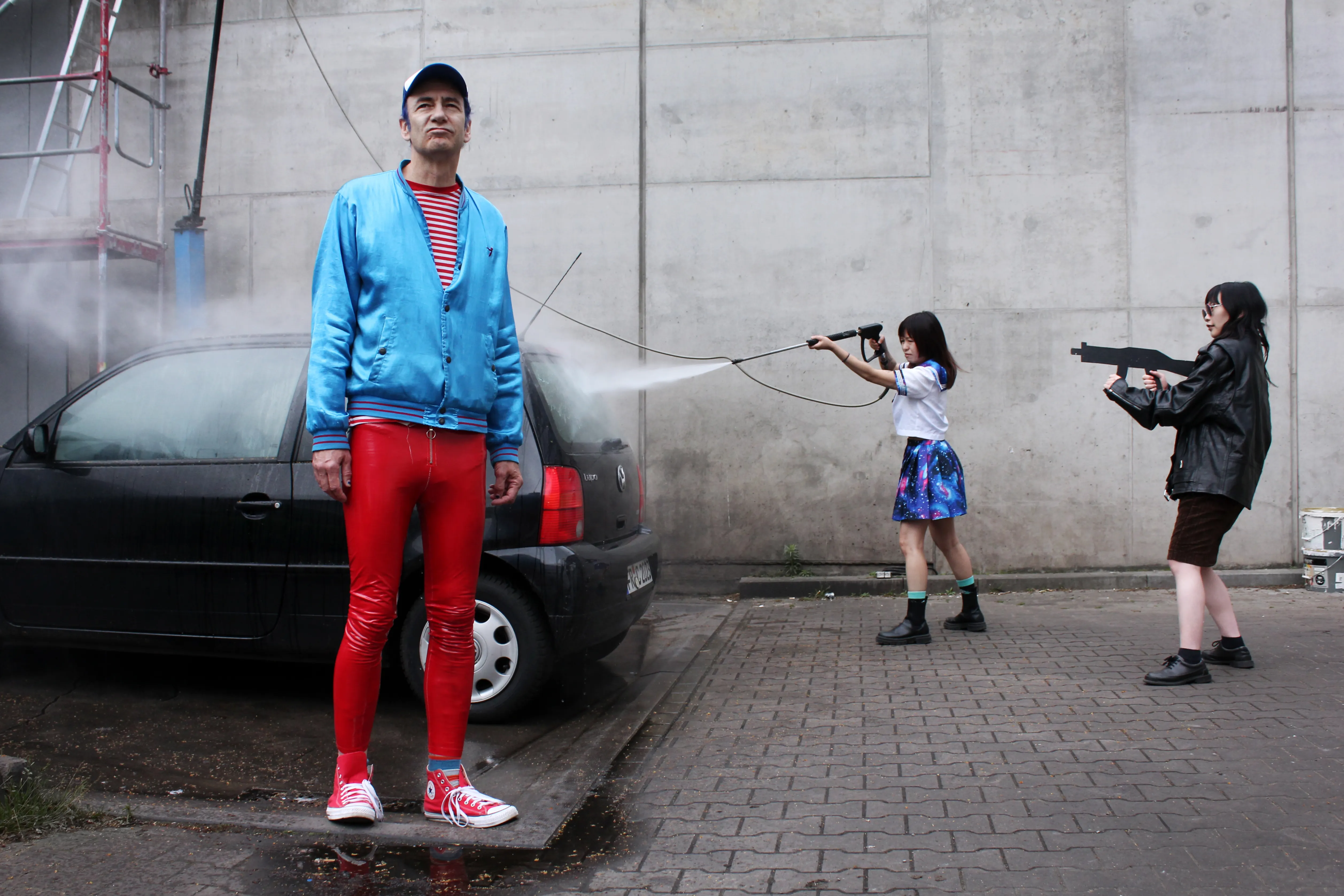 Eine Person steht vor einem schwarzen Auto. Zwei Personen spritzen Wasser auf das Auto.