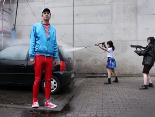 Eine Person steht vor einem schwarzen Auto. Zwei Personen spritzen Wasser auf das Auto.