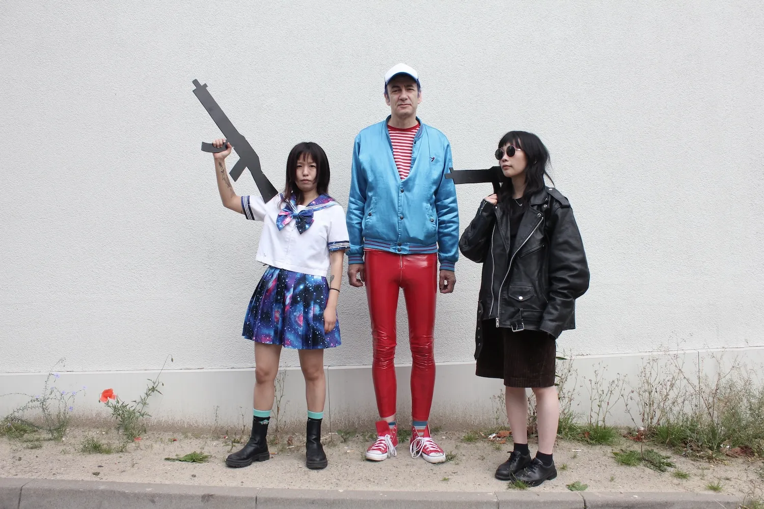 Drei Personen stehen vor einer Wand. Die mittlere in roter Lackhose und einer blauen Jacke, die danebenstehenden halten pistolenähnliche Attrappen in der Hand.