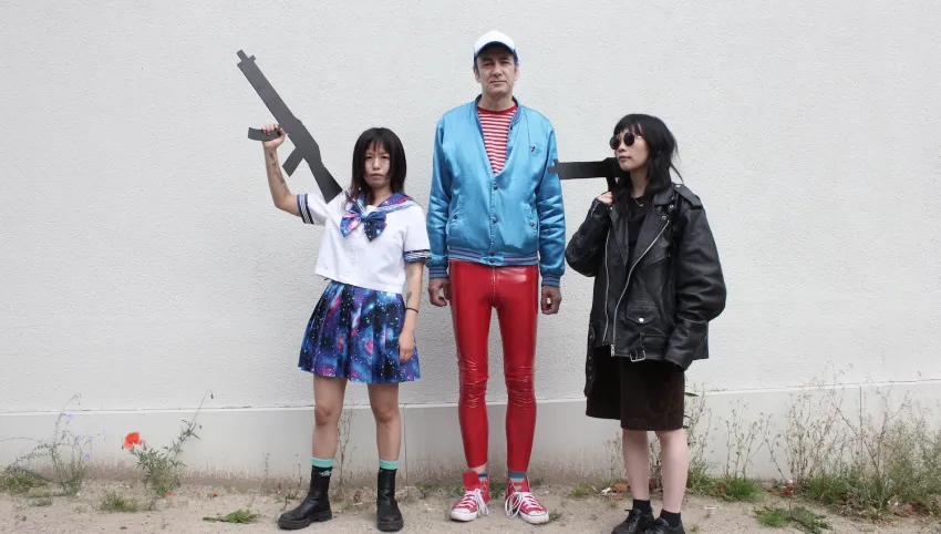Drei Personen stehen vor einer Wand. Die mittlere in roter Lackhose und einer blauen Jacke, die danebenstehenden halten pistolenähnliche Attrappen in der Hand.