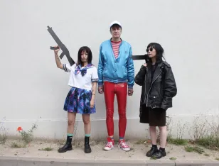 Drei Personen stehen vor einer Wand. Die mittlere in roter Lackhose und einer blauen Jacke, die danebenstehenden halten pistolenähnliche Attrappen in der Hand.