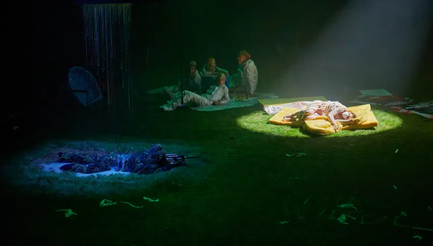 Mehrere Personen liegen auf grünem Boden, wovon eine mit Scheinwerferlicht beleuchtet wird.