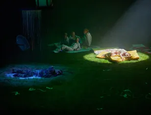 Mehrere Personen liegen auf grünem Boden, wovon eine mit Scheinwerferlicht beleuchtet wird.