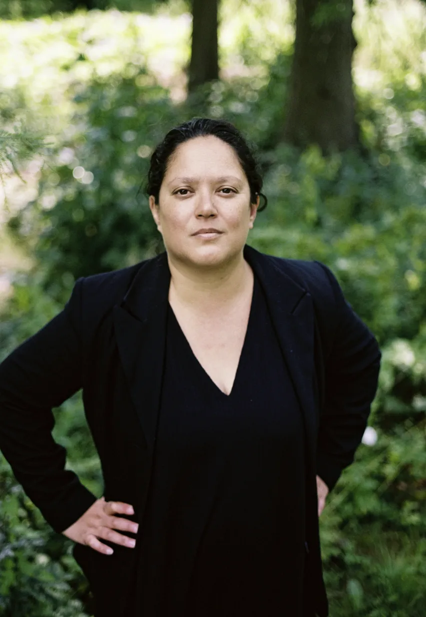 Eine Frau in schwarzer Kleidung, die Hände in die Hüften stemmend. Grüne Natur im Hintergrund.