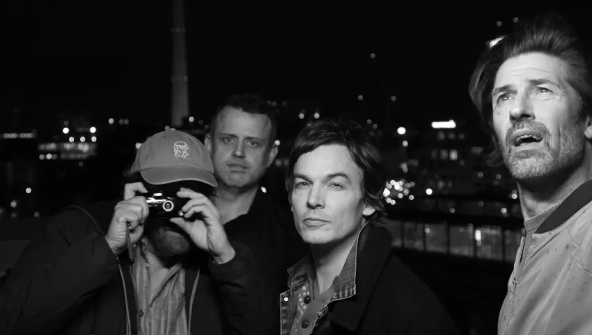 Aufnahme von vier Personen. Die linke hält eine Kamera vor das Gesicht.