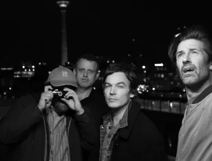 Aufnahme von vier Personen. Die linke hält eine Kamera vor das Gesicht.