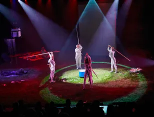 Vier Personen in weißen Anzügen stehen im Kreis und halten lange Stäbe in den Händen.