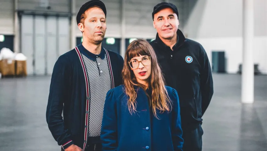 Drei Personen stehen in einer Halle. Im Vordergrund eine Person mit langem Haar und blauem Mantel, hinter ihr zwei, die jeweils eine Kappe tragen.