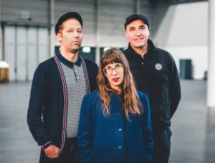 Drei Personen stehen in einer Halle. Im Vordergrund eine Person mit langem Haar und blauem Mantel, hinter ihr zwei, die jeweils eine Kappe tragen.