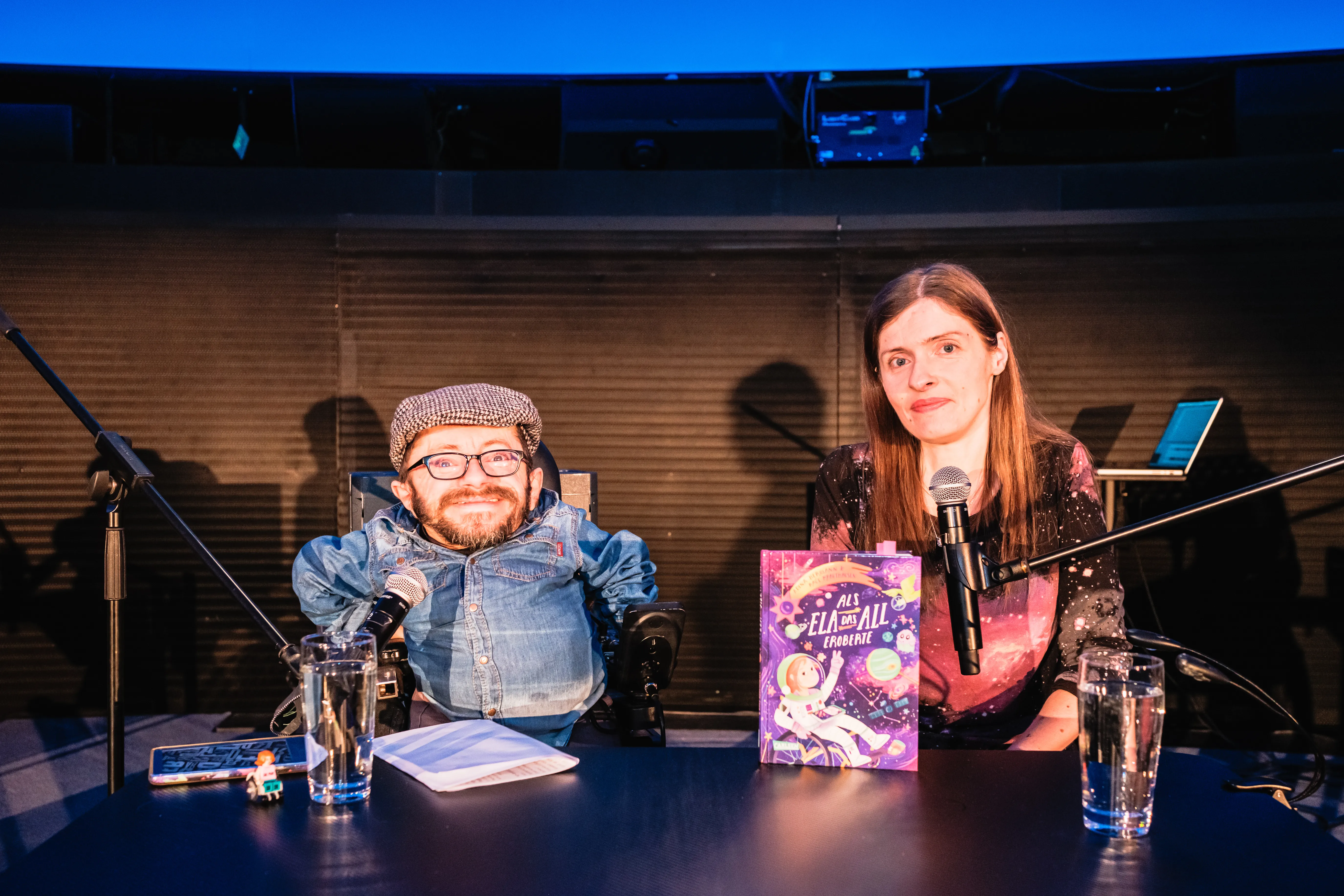 Raúl Krauthausen und Adina Hermann an einem Tisch mit befestigten Mikrofonen sitzend. Auf dem Tisch ihr Buch.