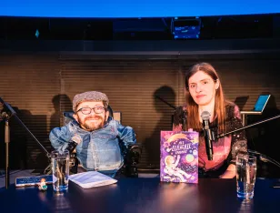 Raúl Krauthausen und Adina Hermann an einem Tisch mit befestigten Mikrofonen sitzend. Auf dem Tisch ihr Buch.