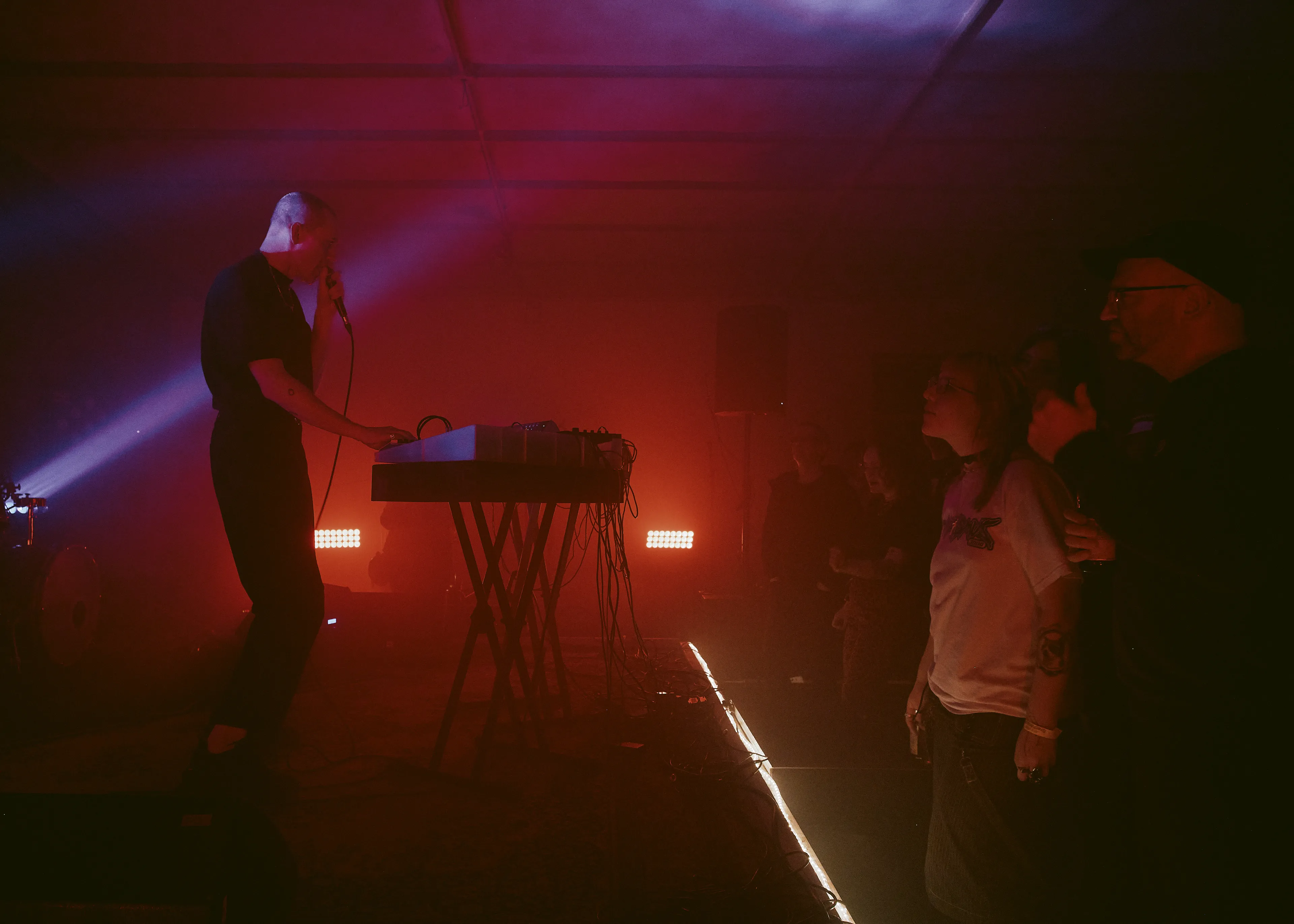 Momentaufnahme vom Konzert: Musiker mit Mikrophon am Synthesizer. Davor stehendes Publikum. Alles in stimmungsvolles rotes Licht getaucht