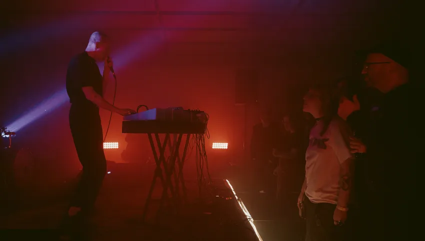 Momentaufnahme vom Konzert: Musiker mit Mikrophon am Synthesizer. Davor stehendes Publikum. Alles in stimmungsvolles rotes Licht getaucht
