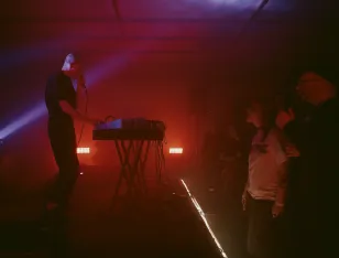 Momentaufnahme vom Konzert: Musiker mit Mikrophon am Synthesizer. Davor stehendes Publikum. Alles in stimmungsvolles rotes Licht getaucht