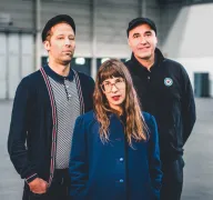 Drei Personen stehen in einer Halle. Im Vordergrund eine Person mit langem Haar und blauem Mantel, hinter ihr zwei, die jeweils eine Kappe tragen.