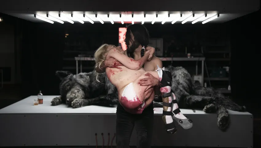 Eine Person, die eine andere, fast unbekleidete Person mit roten Flecken in den Armen hält. Im Hintergrund liegt ein schwarzer Wolf auf einer beleuchteten Fläche.