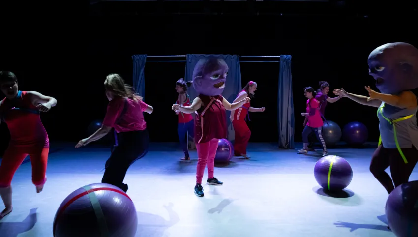 Acht Personen bewegen sich um Gymnastikbälle. Zwei von ihnen haben eine große Maske auf dem Kopf.
