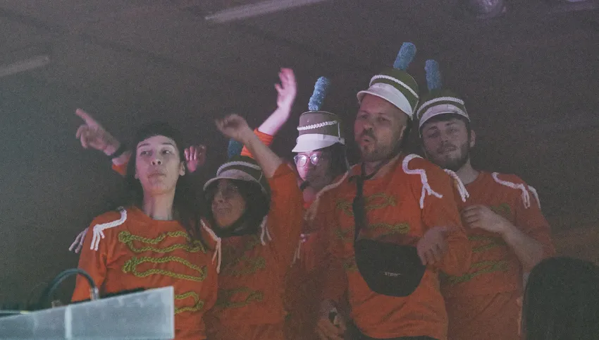 Die Musikong Bumbong Marching Band - eine Gruppe von Künstler*innen in orangefarbenen Uniformen und Hüten tanzt vor der Bühne