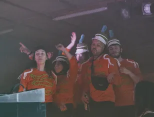 Die Musikong Bumbong Marching Band - eine Gruppe von Künstler*innen in orangefarbenen Uniformen und Hüten tanzt vor der Bühne