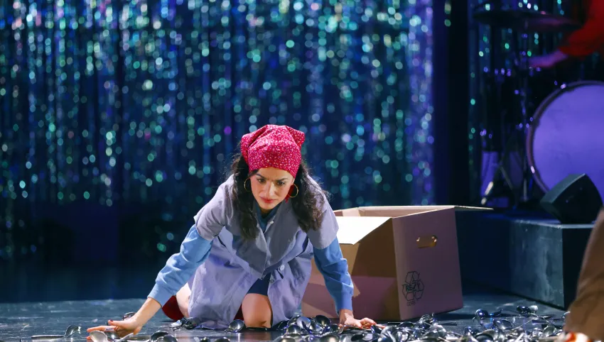 Eine Frau mit roter Kopfbedeckung sitzt vor Löffeln auf dem Boden. Hinter ihr liegt eine offene Kartonkiste.