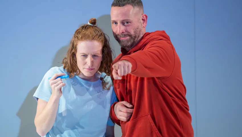 Eine Person trägt ein hellblaues Oberteil, eine andere einen roten Kapuzenpullover und zeigt mit ausgestrecktem Arm nach vorne.