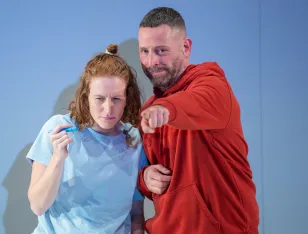 Eine Person trägt ein hellblaues Oberteil, eine andere einen roten Kapuzenpullover und zeigt mit ausgestrecktem Arm nach vorne.