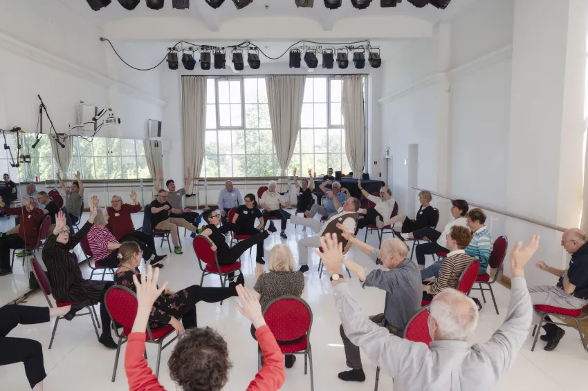 Viele Menschen, die im Stuhlkreis sitzen und ihre Arme und Beine in die Luft halten.