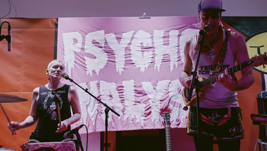 Brezel Göring, ein hochgewachsener Mann mit Baseballcap und Ukulele singt ins Mikro. Rechts hinter ihm  eine Person in schwarzem Achselshirt an den Percussions. Im Hintergrund aufgespannt: ein pinkes Tuch mit der Aufschrift "Psycho-Analyse". In weißen triefenden Großbuchstaben