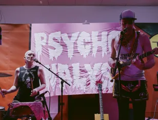 Brezel Göring, ein hochgewachsener Mann mit Baseballcap und Ukulele singt ins Mikro. Rechts hinter ihm  eine Person in schwarzem Achselshirt an den Percussions. Im Hintergrund aufgespannt: ein pinkes Tuch mit der Aufschrift "Psycho-Analyse". In weißen triefenden Großbuchstaben