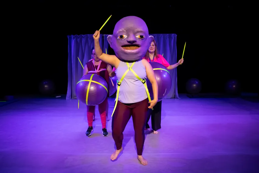 Eine Person mit einer großen Maske auf dem Kopf und einem gelben Stab in der Hand. Hinter ihr stehen zwei weitere Personen mit Gymnastikbällen um ihre Bäuche.