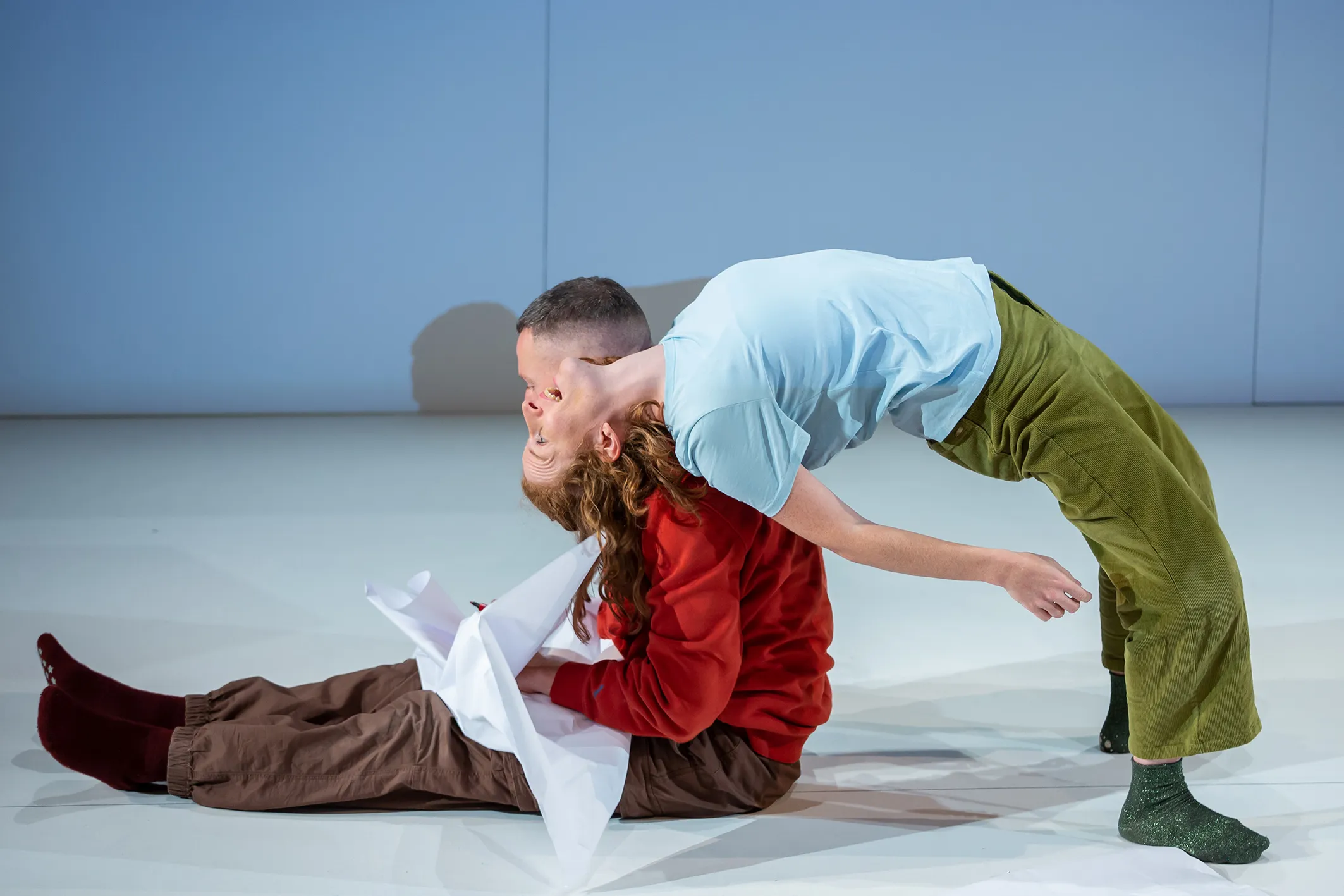 Eine Person sitzt mit brauner Hose und rotem Pullover auf dem Boden, eine andere lehnt mit blauem Oberteil und grüner Hose nach hinten über sie.