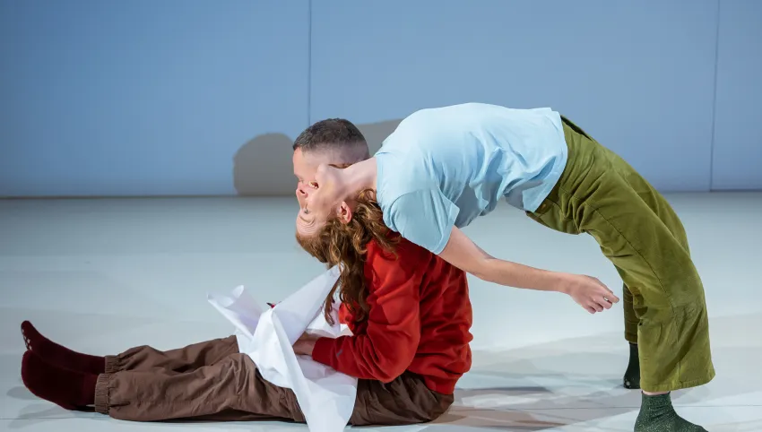 Eine Person sitzt mit brauner Hose und rotem Pullover auf dem Boden, eine andere lehnt mit blauem Oberteil und grüner Hose nach hinten über sie.